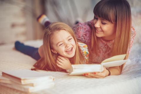 Mother and daughter laugh while reading a book together in bed