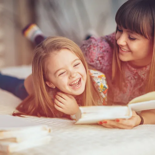 Mother and daughter laugh while reading a book together in bed