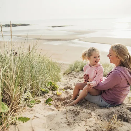 Daughter sits on mother's lap as they relax on a beach.