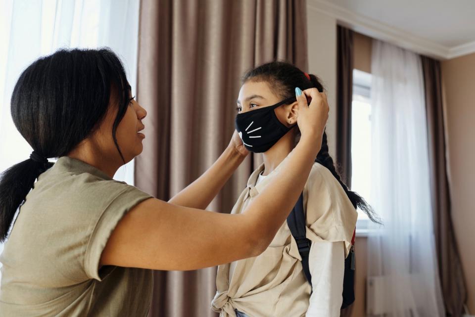 A mother helps her young daughter put on a face mask.